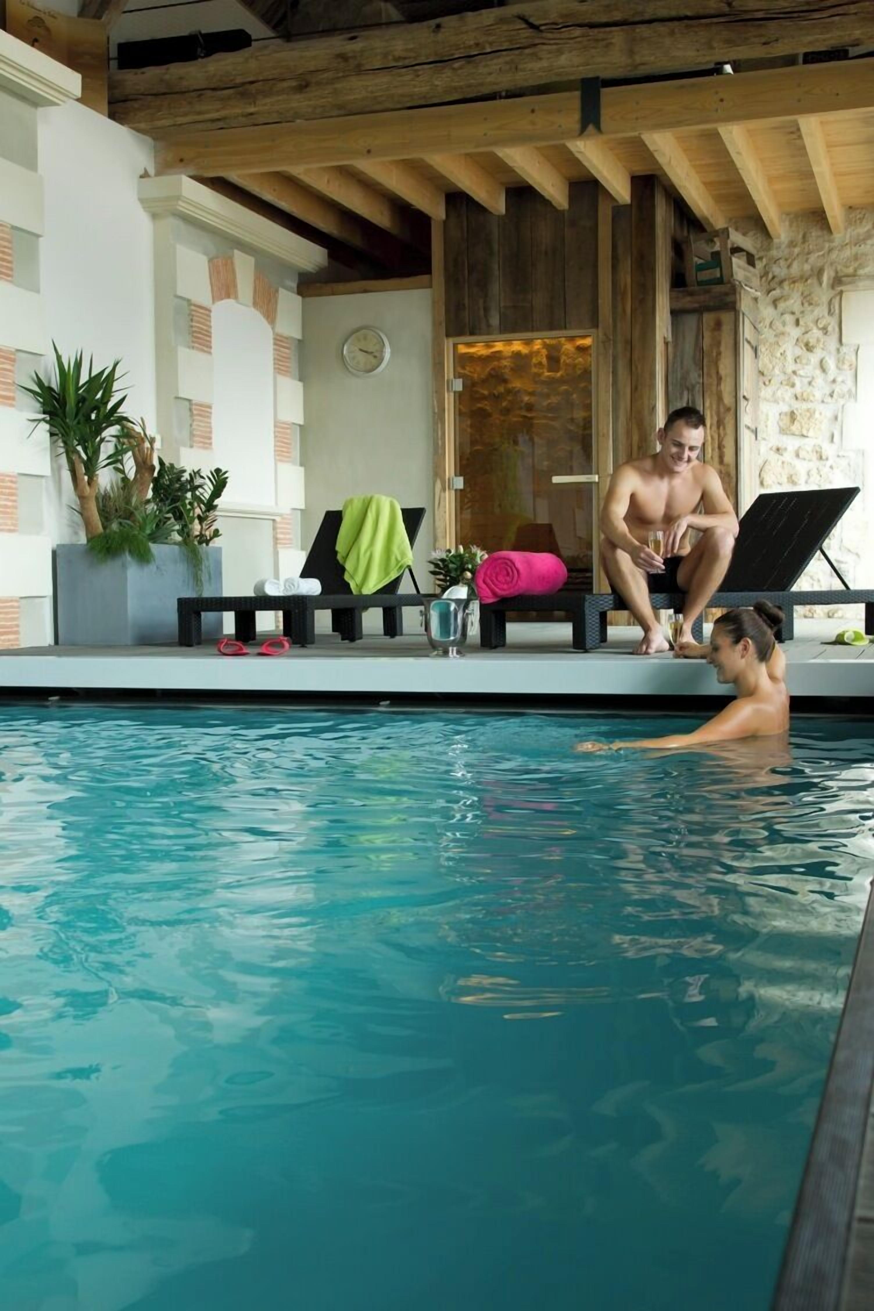 An indoor swimming pool with a wooden ceiling and stone walls. There are lounge chairs and a bench with towels.