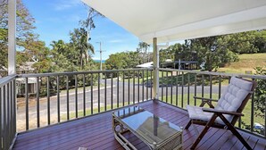 A covered balcony with a glass table and a wooden chair, overlooking a road and greenery.