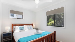 A neatly arranged bedroom with a wooden bed, two bedside tables, a nightstand, a large window with a view of greenery, and a ceiling fan.