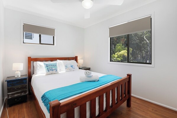 A neatly arranged bedroom with a wooden bed, two bedside tables, a nightstand, a large window with a view of greenery, and a ceiling fan.