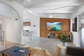 A modern living room with a wooden accent wall, a flat-screen TV, and a dining area with white chairs. - Welcome to Malibu - Your Laid-Back Straddie Beach Getaway (Point Lookout)