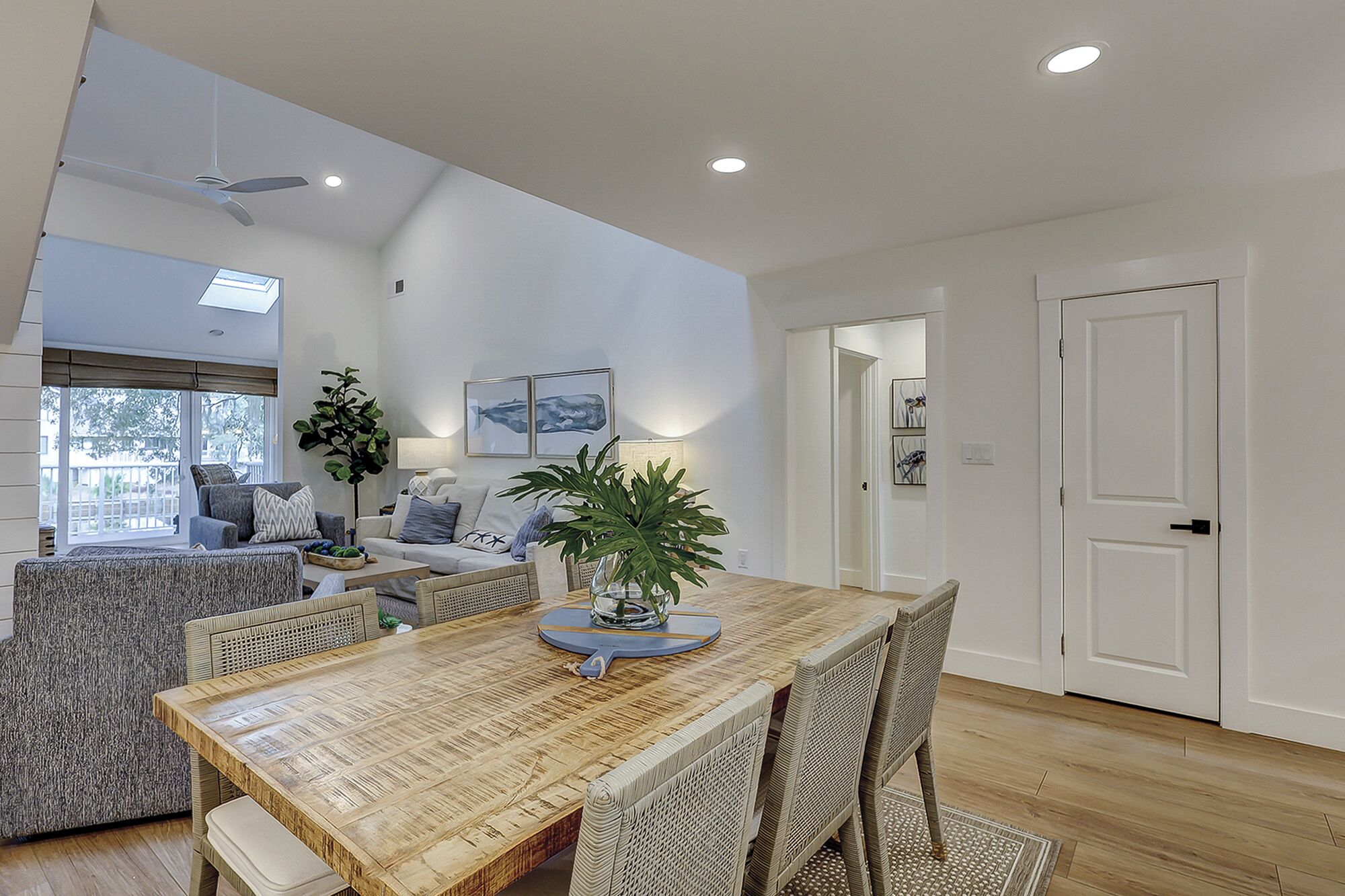 Un coin repas avec une table en bois, des chaises en osier et une plante en pot. On y trouve un ventilateur de plafond, un éclairage encastré et une vue sur l’extérieur par une fenêtre.