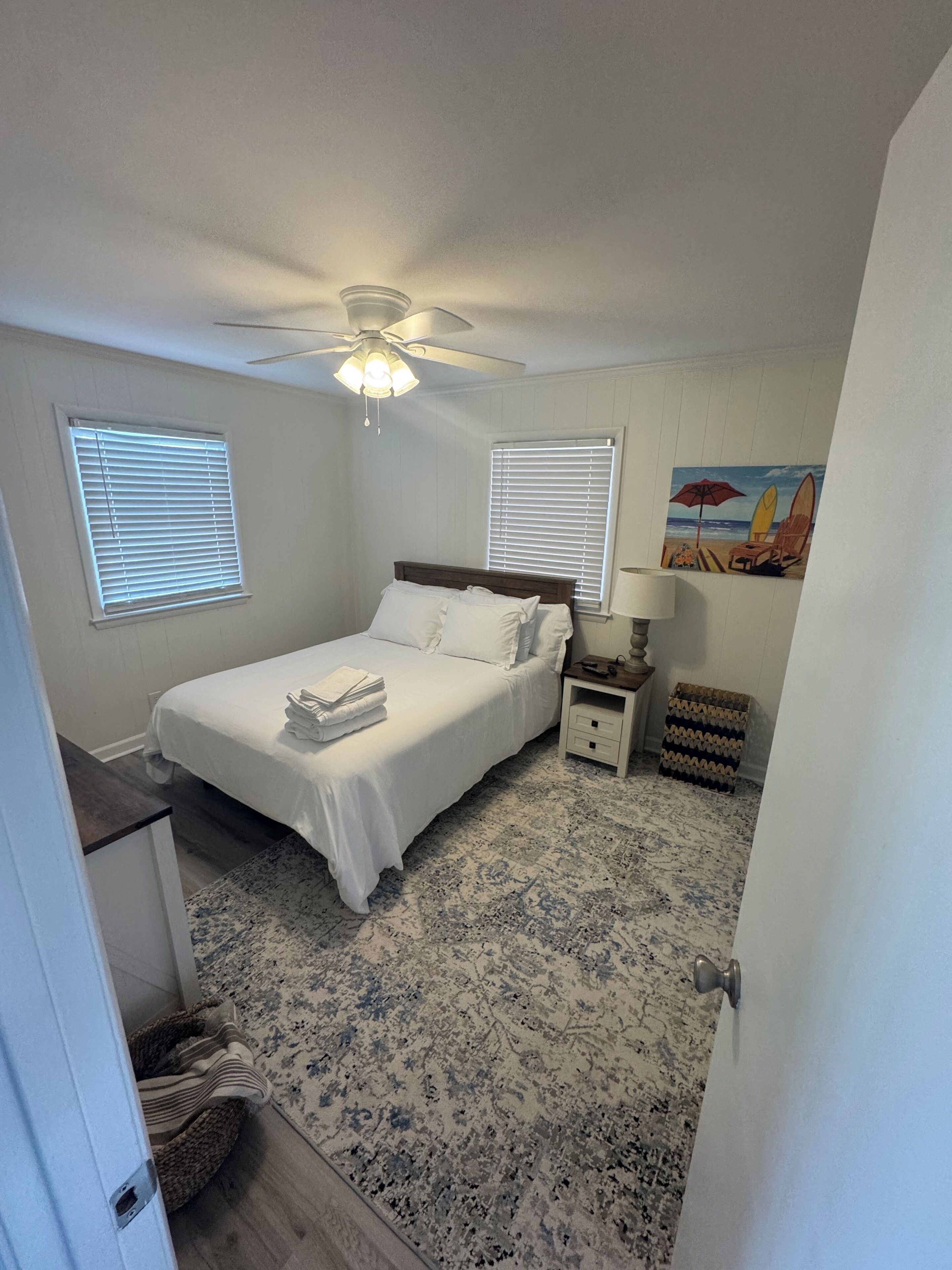 A bedroom with a bed, two bedside tables, a ceiling fan, and a beach-themed painting.