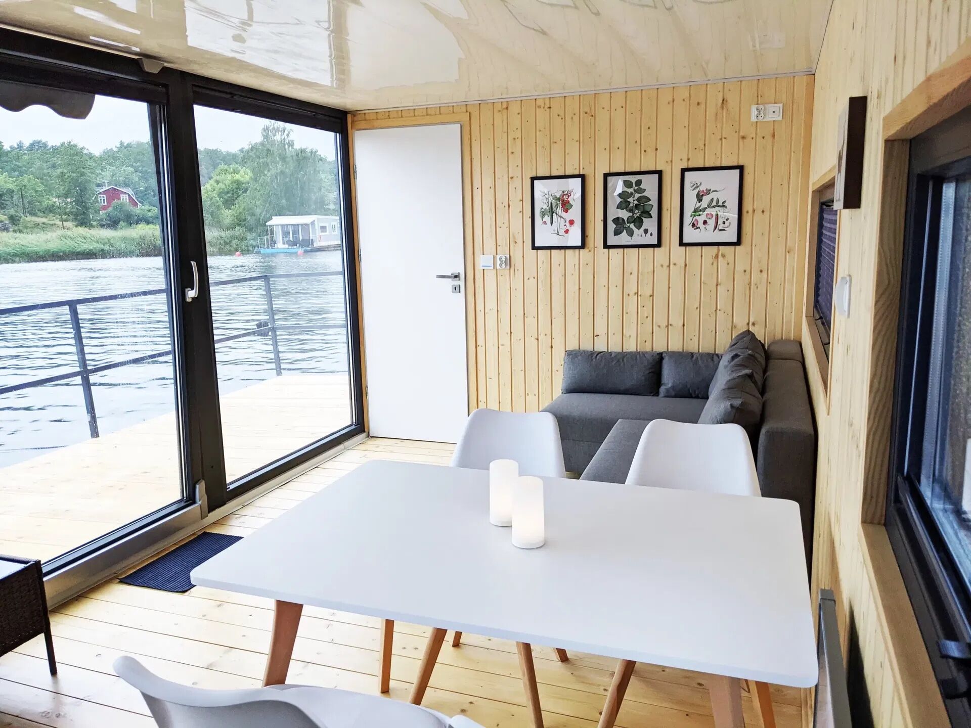 A cabin with a wooden interior, a white table with candles, a sofa with cushions, and a view of a river and buildings through large windows.