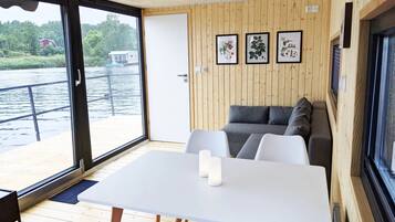 A cabin with a wooden interior, a white table with candles, a sofa with cushions, and a view of a river and buildings through large windows.
