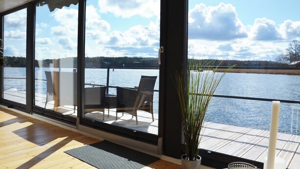 A room with large windows overlooking a body of water, a wooden floor, and a seating area with chairs and a table. - The Saltkrokan Houseboat in the Archipelago of Southern Sweden (Blankaholm)