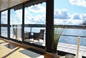 Een kamer met grote ramen met uitzicht op een wateroppervlak, een houten vloer en een zithoek met stoelen en een tafel.