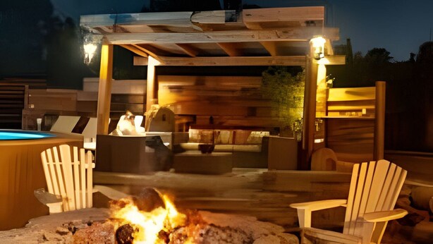 Un hogar de piedra con fuego, rodeado de sillas Adirondack, bajo una pérgola de madera con asientos.