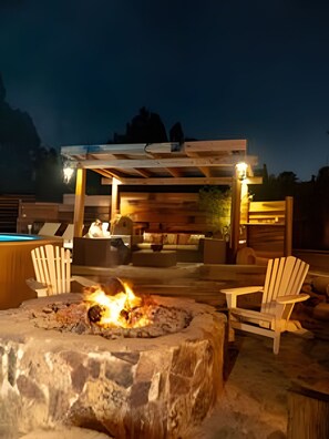 A stone fire pit with a fire, surrounded by Adirondack chairs, under a wooden pergola with seating. - Oceana Suites Parcela San Ramon (La Serena)