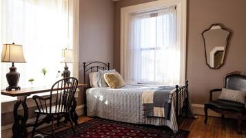 Ein Schlafzimmer mit Bett, Schreibtisch, zwei Stühlen, einem Fenster mit leichtem Vorhang und einem Spiegel an der Wand.