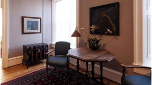 Une pièce avec une table en bois, une chaise bleue, une valise ancienne, une lampe et un tableau accroché au mur.
