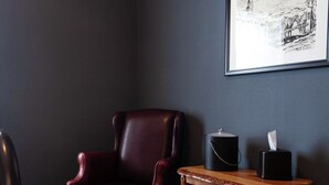A corner room with a dark wall, a wooden chair, a small wooden table with a tissue box and a black container. - Fred Lambert - Historic Hotel Room (Cimarron)