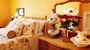 Une chambre à coucher avec une couvre-lit à motifs floraux, une lampe de chevet, un miroir et un vase de fleurs posé sur une commode en bois.
