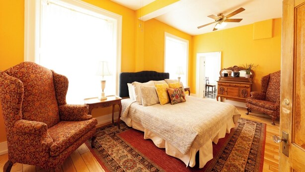 A bedroom with a bed, two chairs, a nightstand, a dresser, and a ceiling fan. - William F. Cody - Historic Hotel Room (Cimarron)