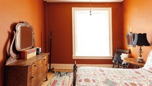 A bedroom with a large window, a dresser, a mirror, a lamp, and a bed with a floral bedspread. - Zane Grey - Historic Hotel Room (Cimarron)