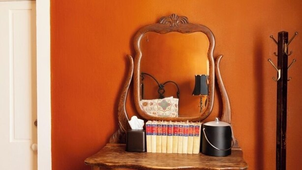 A wooden dresser with a mirror, books, a tissue box, and a lamp. - Zane Grey - Historic Hotel Room (Cimarron)