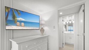 A modern bathroom with a large mirror, white cabinetry, and a flat-screen TV displaying a beach scene. - Sundial A203 - Stunning, Gulf Front, 1 bedroom Condo on Sanibel Island! (Sanibel)