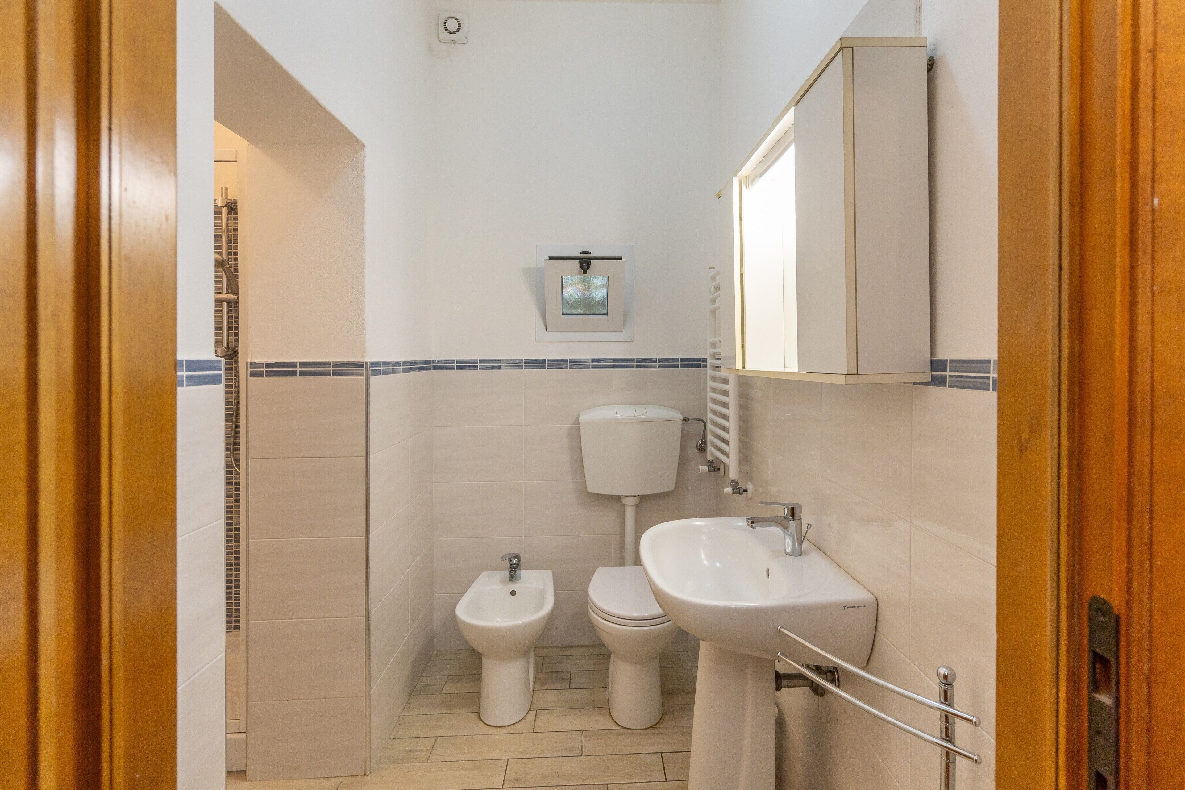 Een badkamer met toilet, bidet en wastafel, betegelde wanden en een houten deurkozijn.
