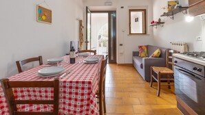 Een eethoek met geruit tafelkleed, stoelen, een eettafel, een bank, een keuken met fornuis, en een deur naar een buitenruimte.