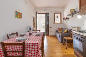 A dining area with a checkered tablecloth, chairs, a dining table, a sofa, a kitchen with a stove, and a door leading to an outdoor area. - Apartment 'Via Gorizia 8 - Rosso' with Private Garden and Wi-Fi (Cecina)