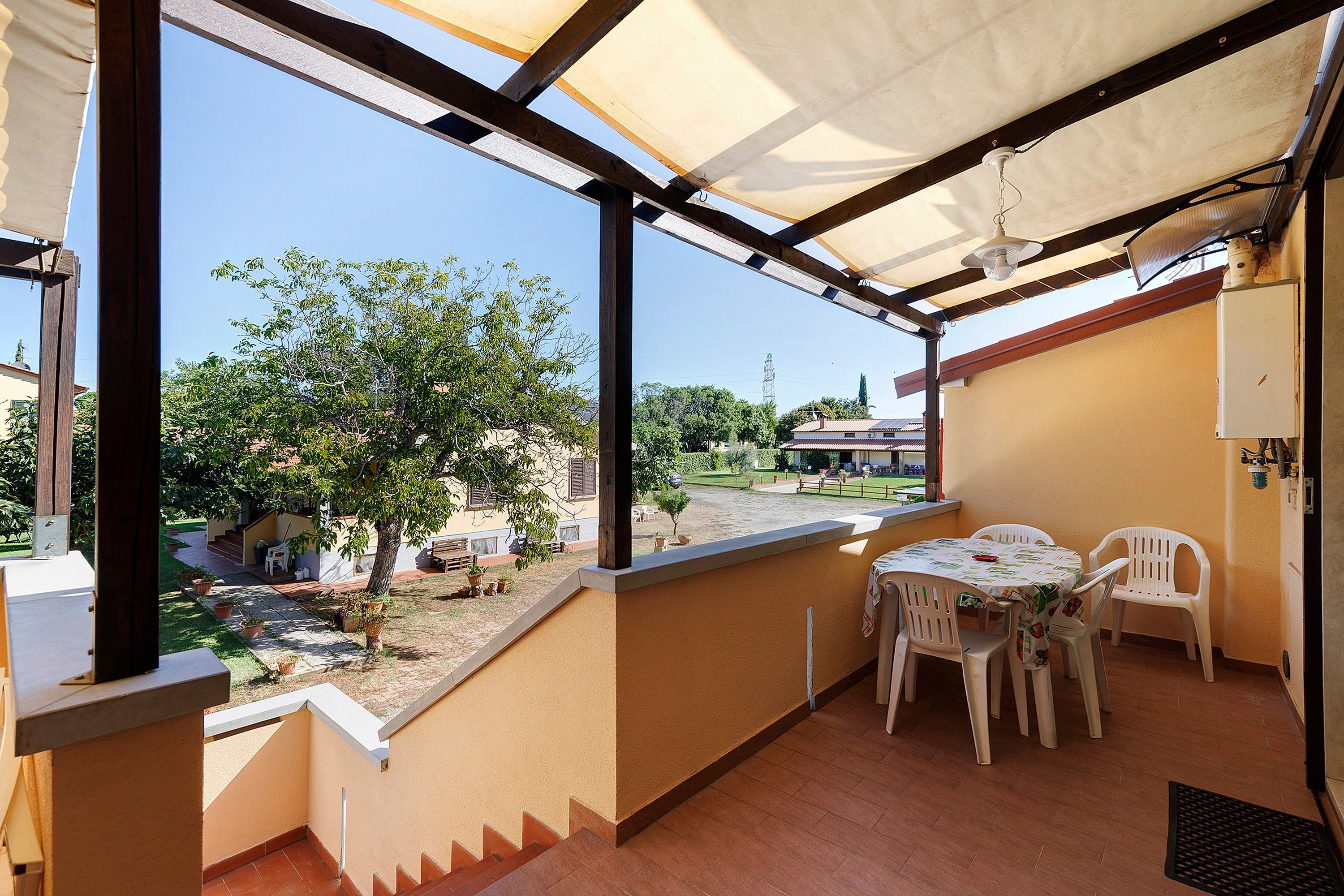 A balcony with a table and chairs, a view of a garden, and a building in the background.