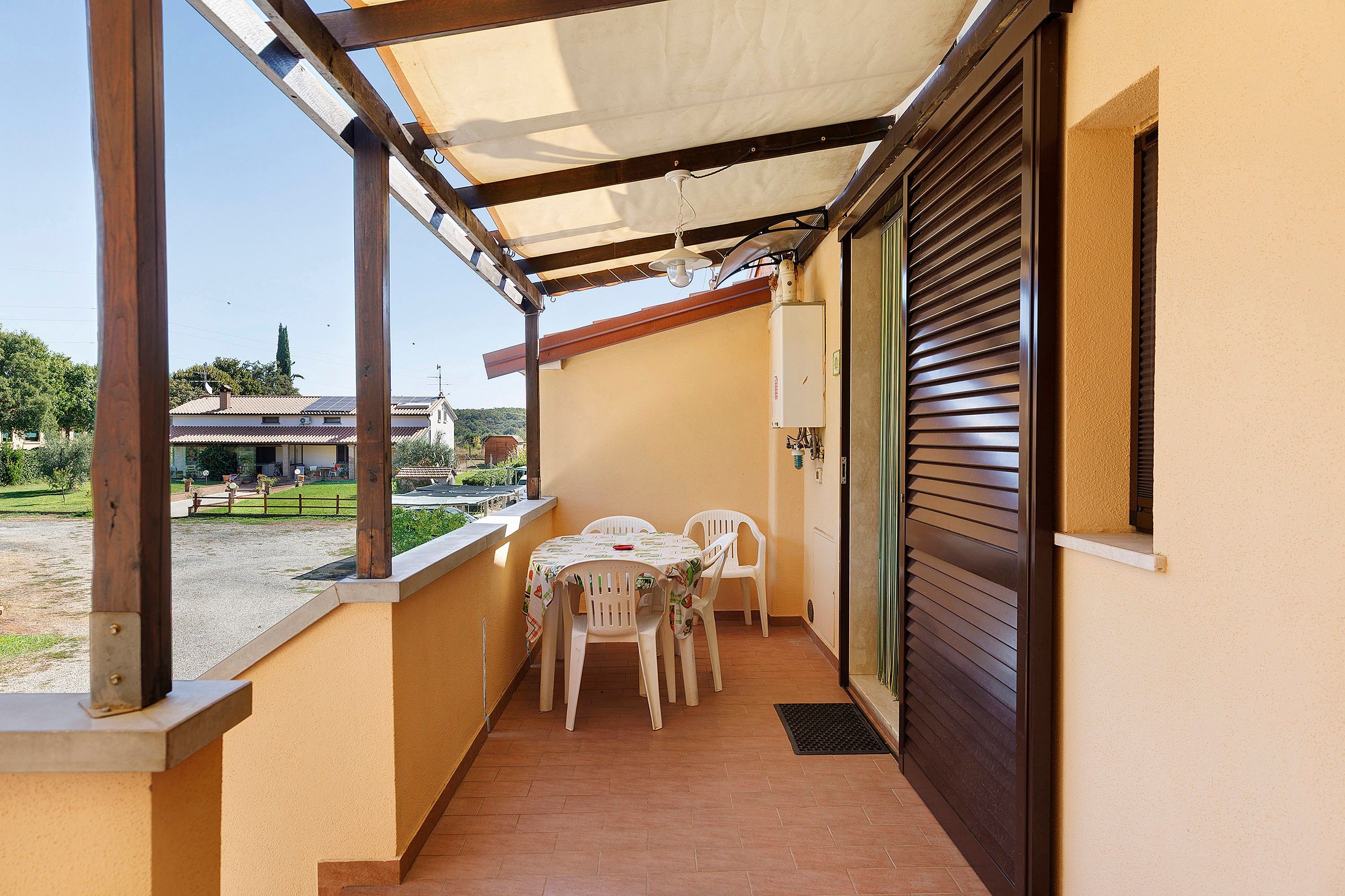 Veranda esterna coperta con tavolo e sedie, vista sulla strada e un edificio con persiana chiusa.