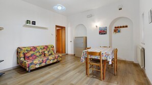 A room with a wooden floor, a floral sofa, a dining table with chairs, and a ceiling light fixture. - Apartment 'Via Gorizia 8 - Verde' with Shared Garden, Wi-Fi and Air Conditioning (Cecina)