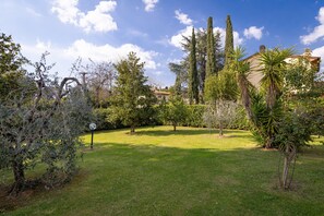 Un jardí ben cuidat amb diversos arbres i arbustos, un prat i un edifici al fons.