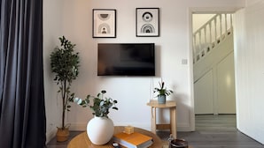 A living room with a flat-screen TV, two framed artworks, a potted plant, a book, and a small table.