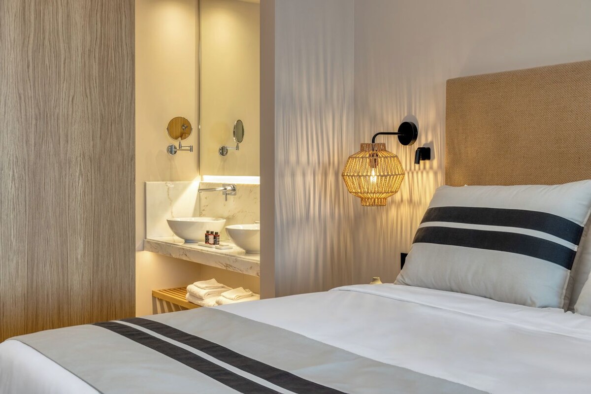 Une chambre d’hôtel comprenant un lit, une table de chevet, un miroir, un luminaire fixé au mur et une salle de bain avec un lavabo et un miroir.