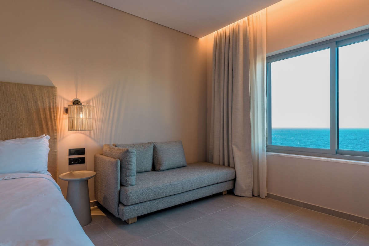 Una habitación de hotel moderna con una cama, un sofá, una mesita de noche, una lámpara y vistas al mar a través de un ventanal grande.