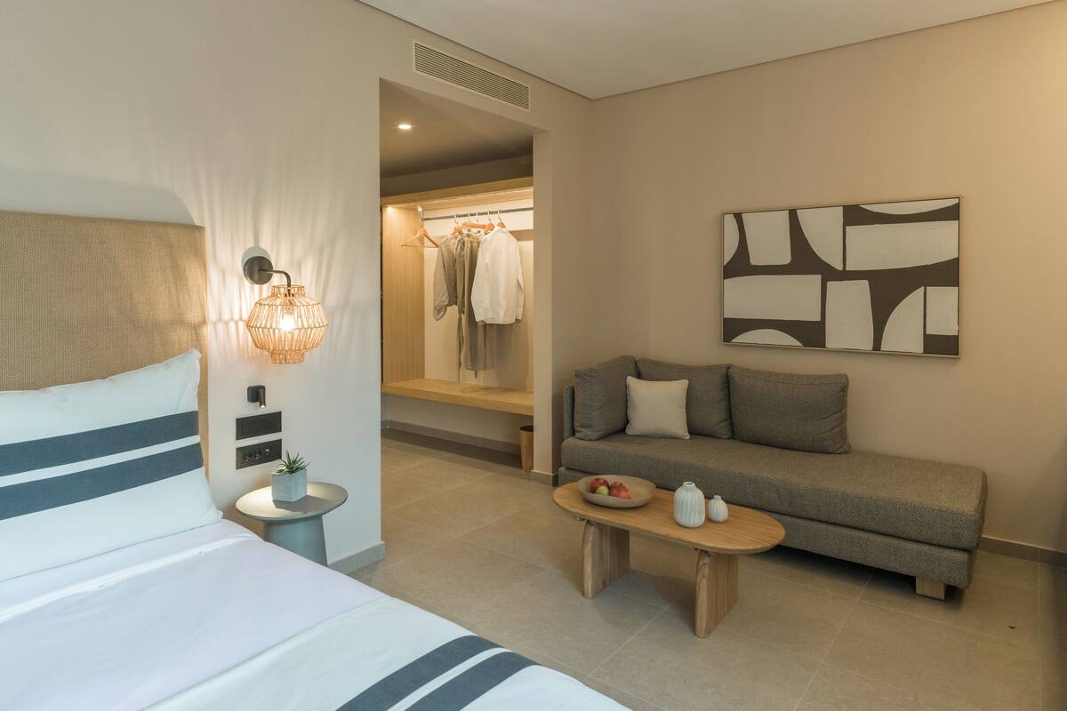 Una habitación de hotel con una cama, un sofá, una mesita con un frutero, una lámpara de pared y un perchero.