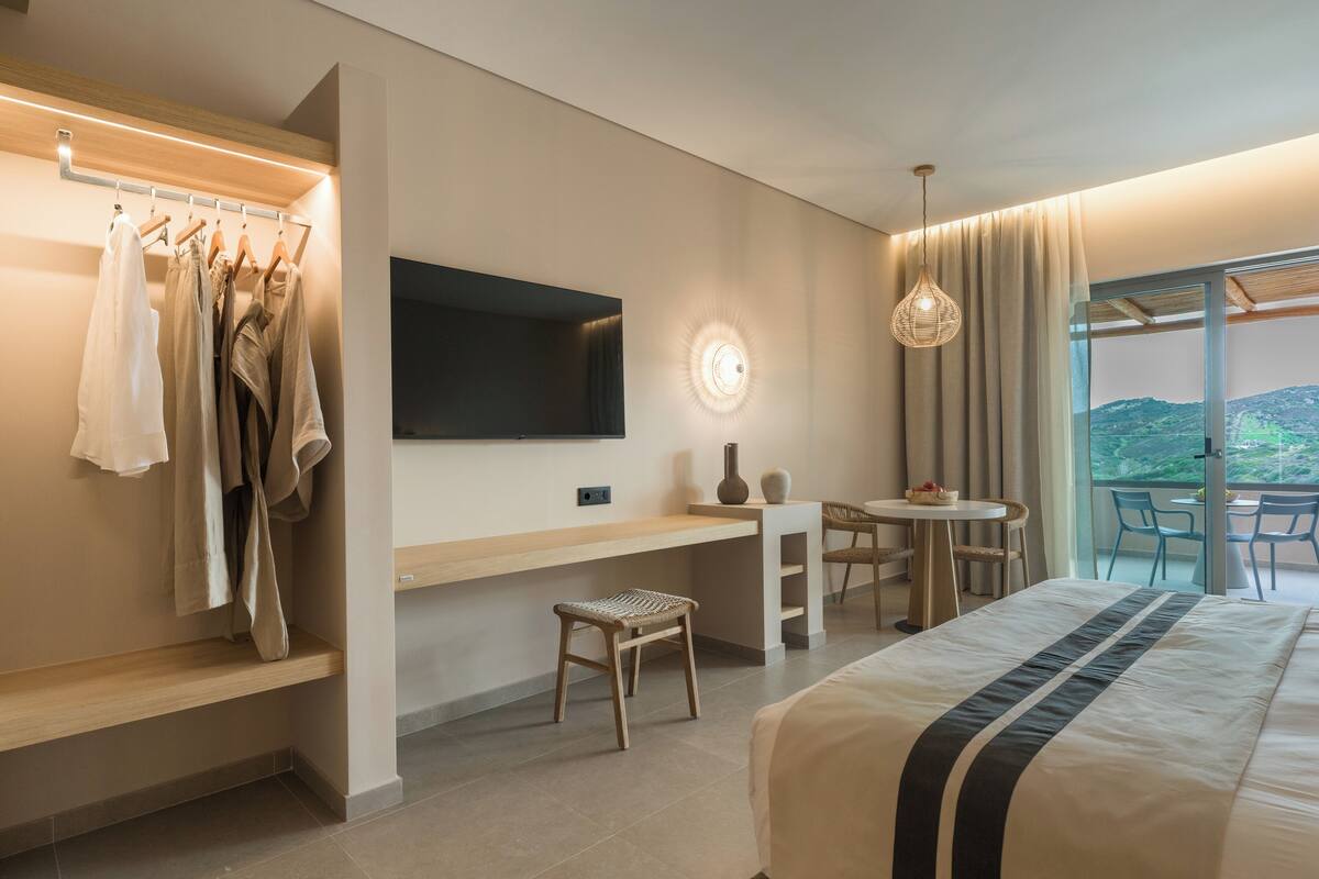 Habitación de hotel con cama, escritorio, silla, televisor, mesita con frutero y vistas al exterior.