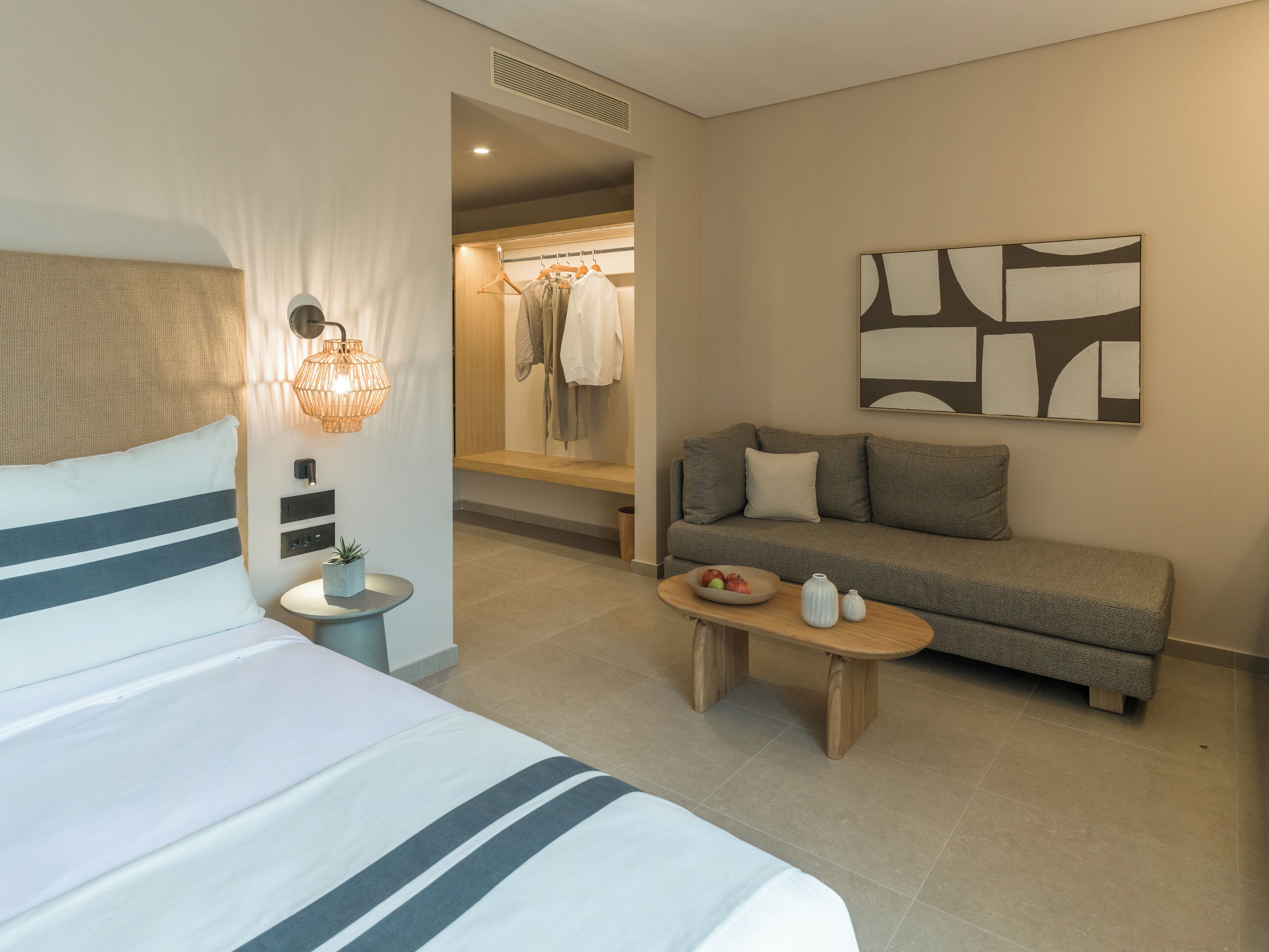 Une chambre d’hôtel avec un lit, un canapé, une petite table sur laquelle se trouve un saladier de fruits, un luminaire fixé au mur et un tableau.