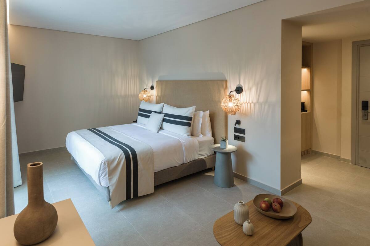 Una habitación de hotel moderna con una cama grande, lámparas de noche, una mesita redonda con un frutero y un florero.