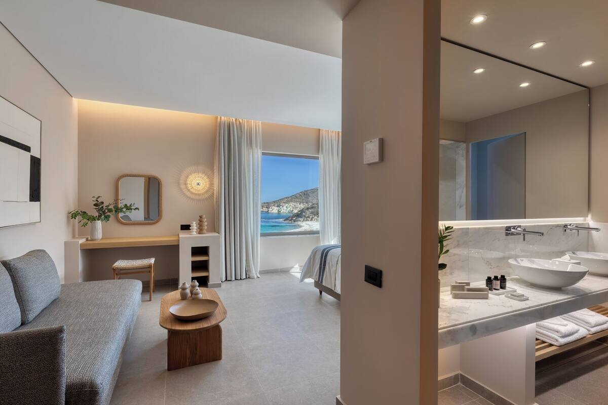 Una habitación de hotel moderna con cama, zona de estar, baño con espejo grande y lavamanos, y vista al mar.