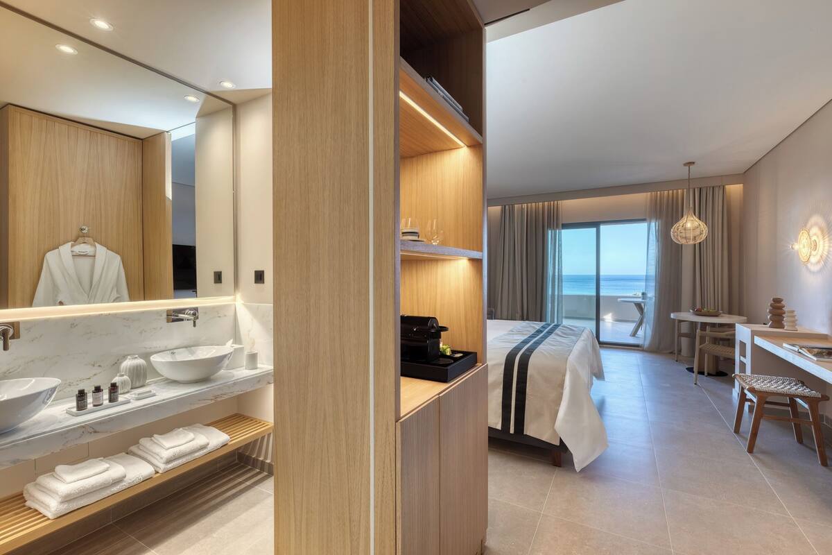 Una habitación de hotel moderna con un espejo grande, un baño con lavamanos y toallero, una cama con vista al mar, un comedor con mesa y sillas, y un balcón con vistas.