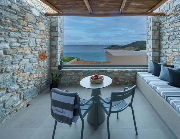 Un balcón de paredes de piedra con vistas al mar, una mesa redonda con un frutero y un sofá con cojines.