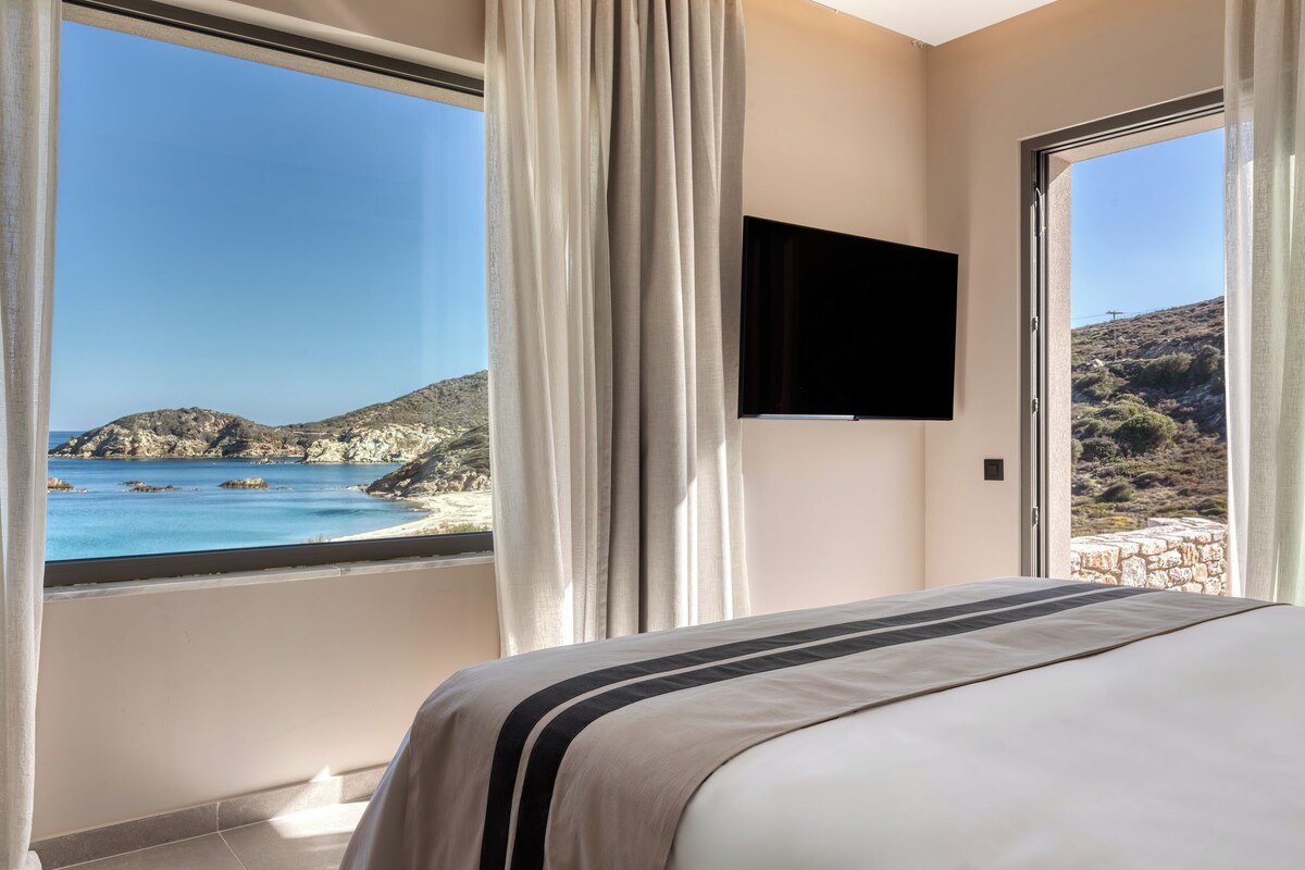 Een slaapkamer met een groot raam met uitzicht op een strand en rotsachtig landschap.