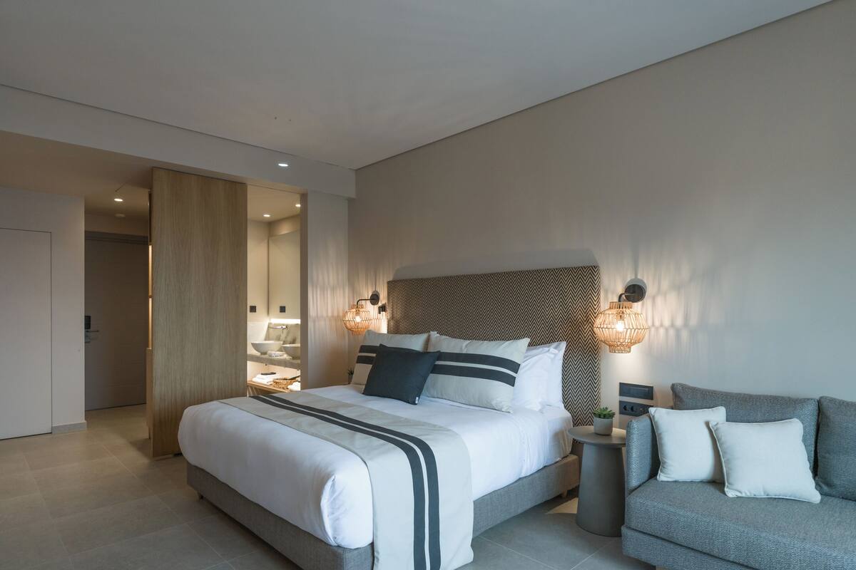 Una habitación de hotel moderna con una cama grande, mesitas de noche, un sofá y un baño visible a través de una puerta abierta.