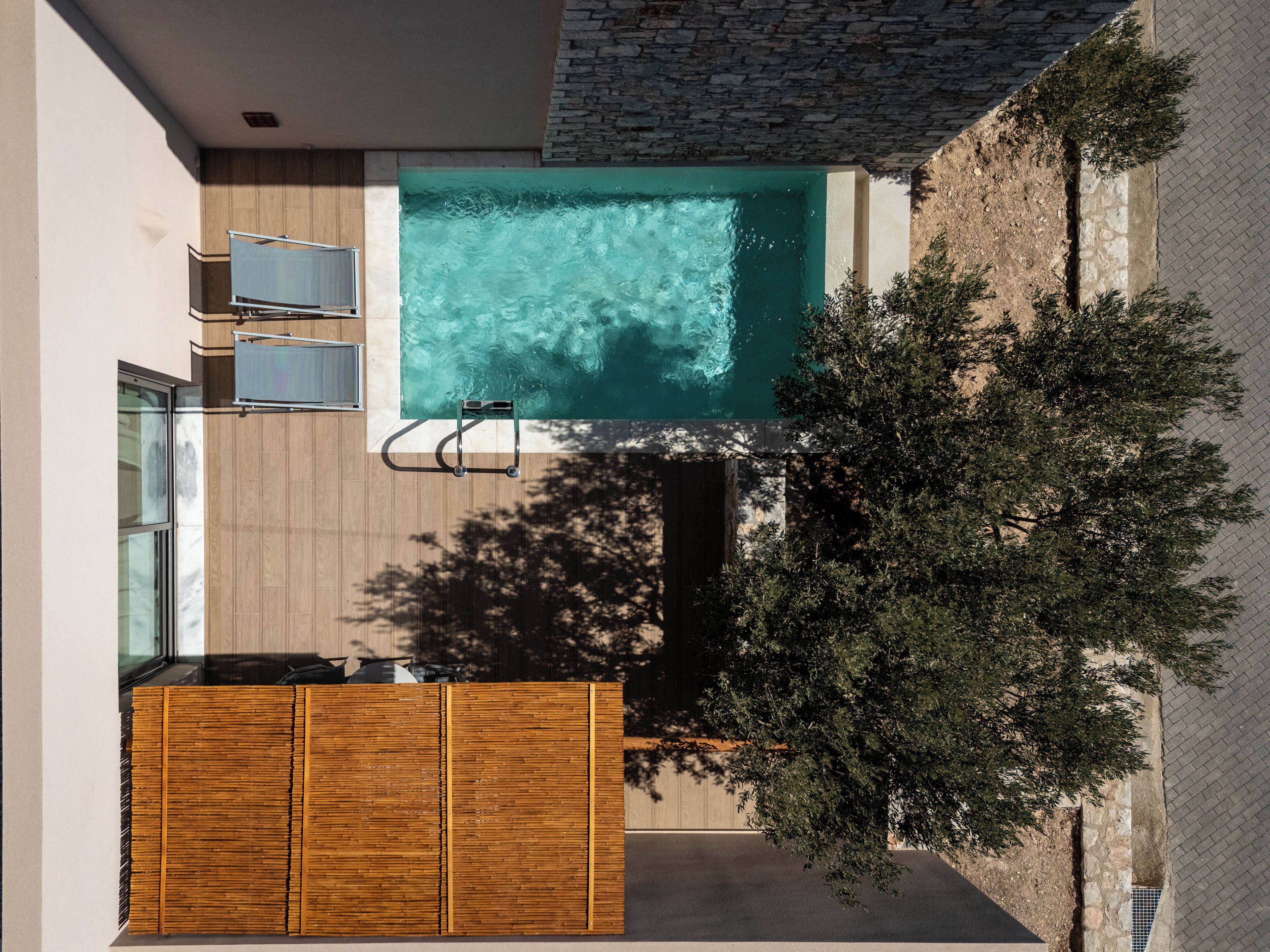 Una piscina exterior moderna con una terraza de madera, dos sillones de descanso y un árbol.