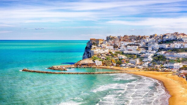 Une ville côtière dotée d’une plage de sable, d’une digue rocheuse et d’une colline densément bâtie.