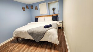 Una cama bien hecha con cabecera de madera, una mesita de noche con lámpara y una ventana con persianas.