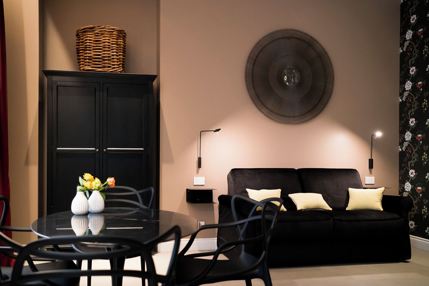 Un salon moderne avec un canapé noir, deux tables d’appoint avec des lampes, un élément décoratif rond fixé au mur, un meuble noir et une table à manger avec des chaises.