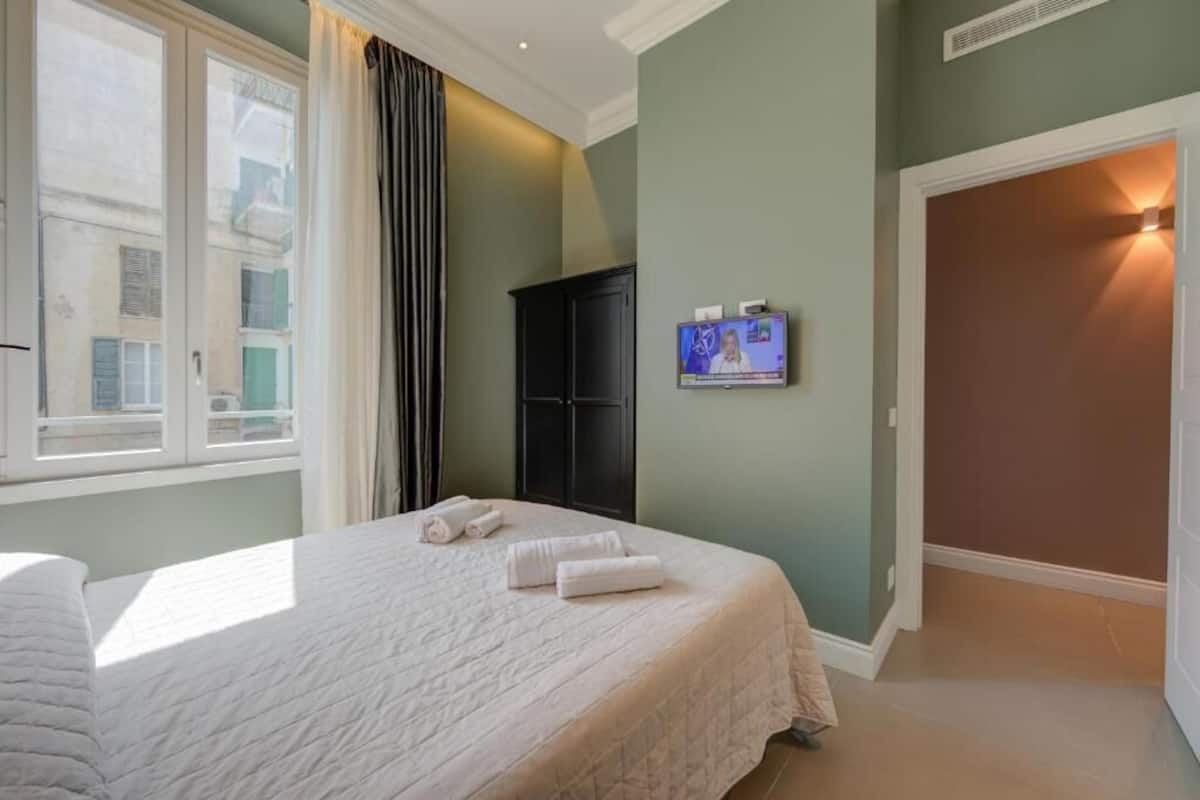 A hotel room with a bed, a TV on the wall, and a window with curtains.