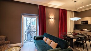 A living room with a dark green sofa, a dining table with chairs, a kitchen area, and a view of a city street through a window. - SEVEN SUITES APARTMENTS SANREMO (Sanremo)