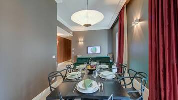 Un comedor con una mesa oscura dispuesta para seis personas, rodeada de sillas, un sofá verde y un televisor montado en la pared.