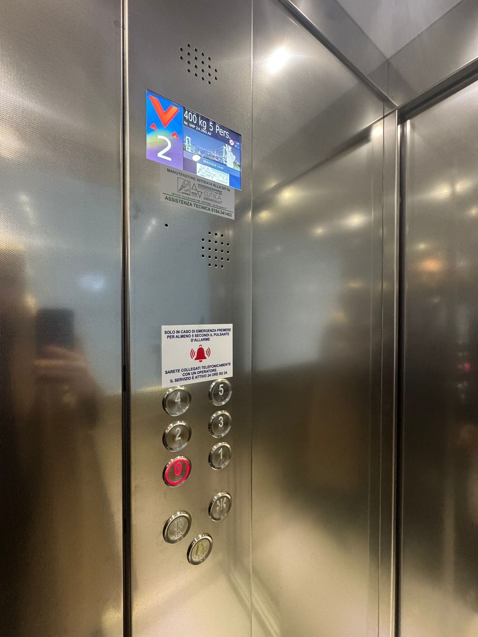 Panel de control de ascensor con pantalla digital, instrucciones de emergencia y número de teléfono para asistencia técnica.