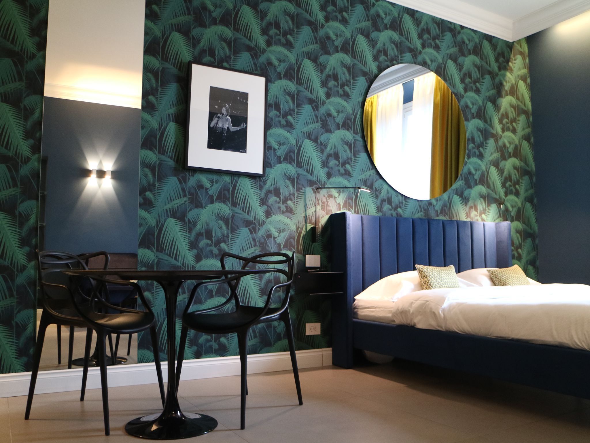 Una camera da letto con un letto, uno specchio rotondo, un quadro incorniciato, un tavolo con sedie e una parete decorata con un motivo a foglia tropicale.