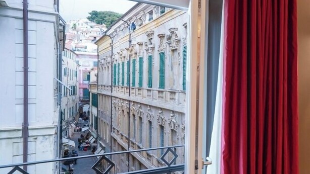 A balcony with a view of a narrow street lined with buildings, a metal railing, and a red curtain. - SEVEN SUITES APARTMENTS SANREMO (Sanremo)